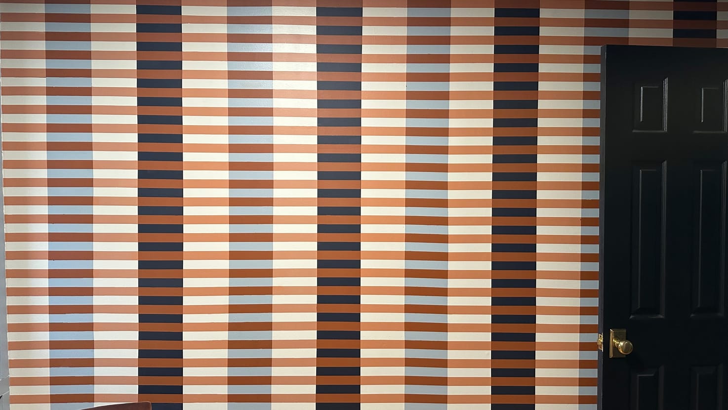 Striped wall art with vertical bands in brown, orange, cream, and blue creates a retro feel. A black door is on the right, and part of a brown chair is visible on the left.