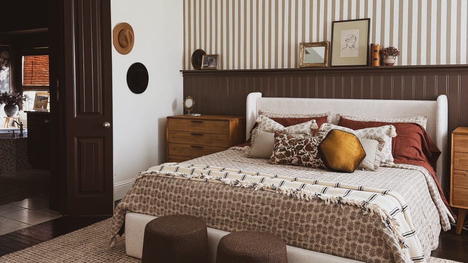 Cozy bedroom with a queen bed, patterned bedding, and warm-toned pillows. Two brown ottomans at the foot, striped accent wall, and wooden nightstands.
