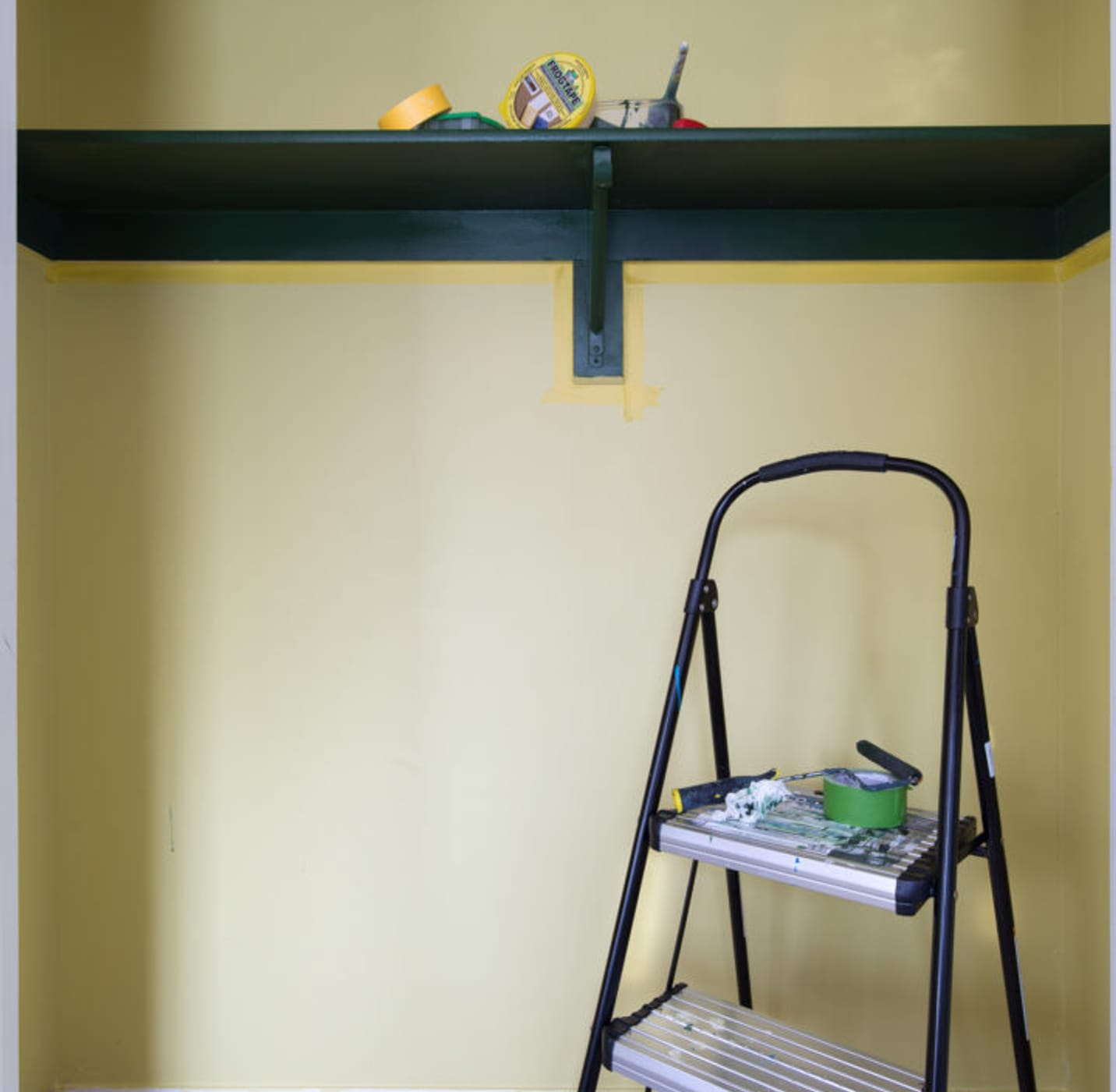 Beige wall with long brown shelf hung near the top. There is a step ladder and the floor is covered by a drop cloth