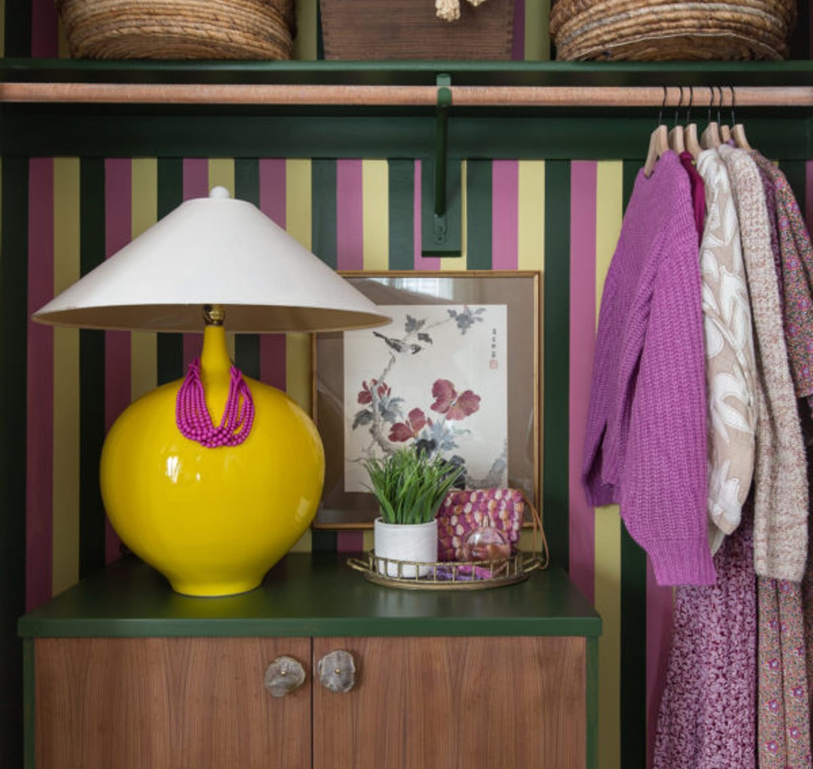Pink, yellow, and green striped closet. There is a small vanity with a yellow lamp, succulent, and frame, with a few clothes hanging in the closet.