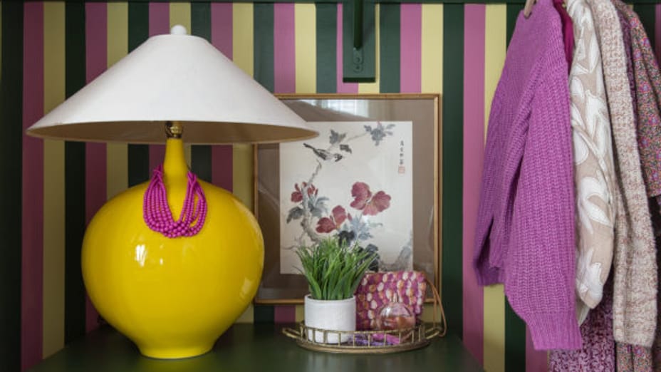 Pink, yellow, and green striped closet. There is a small vanity with a yellow lamp, succulent, and frame, with a few clothes hanging in the closet.