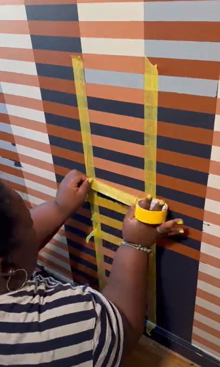A person applies yellow tape on a striped wall with horizontal orange and blue-gray lines. The person wears a striped shirt and bracelet, focusing intently.