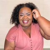 Smiling woman in a pink top and blue jeans stands against a light brick wall. She has curly hair and wears hoop earrings, conveying a cheerful mood.
