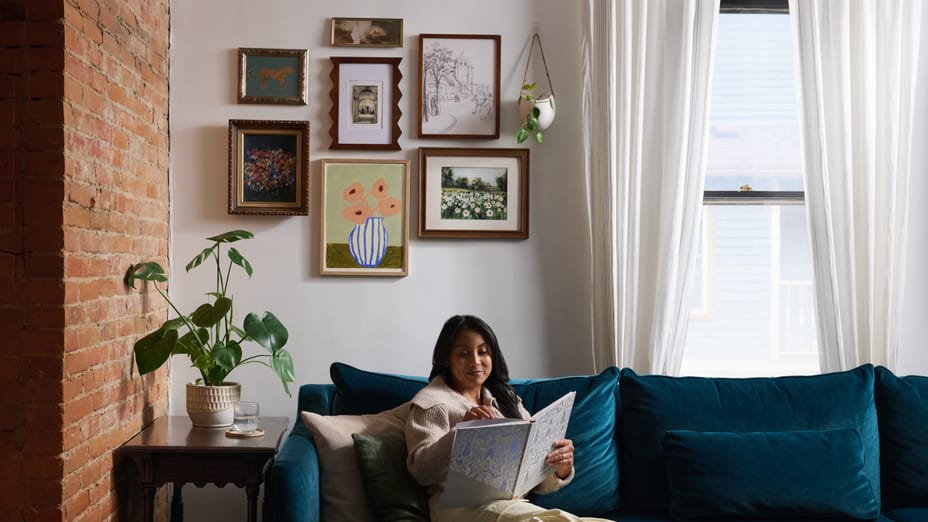 Woman reading a book on a blue couch, behind her are pictures hung on the wall with FrogTape reMOVEables products