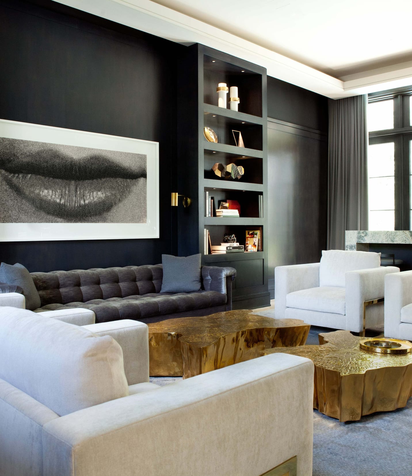 Trendy living room space with black walls, four white chairs, and a dark leather couch with cold colored coffee tables in the middle. The back wall has two book shelves, one on the right and one on the left side of the couch. On the wall in-between is a pint of lips and there is a small bar in front of two windows on the back right wall