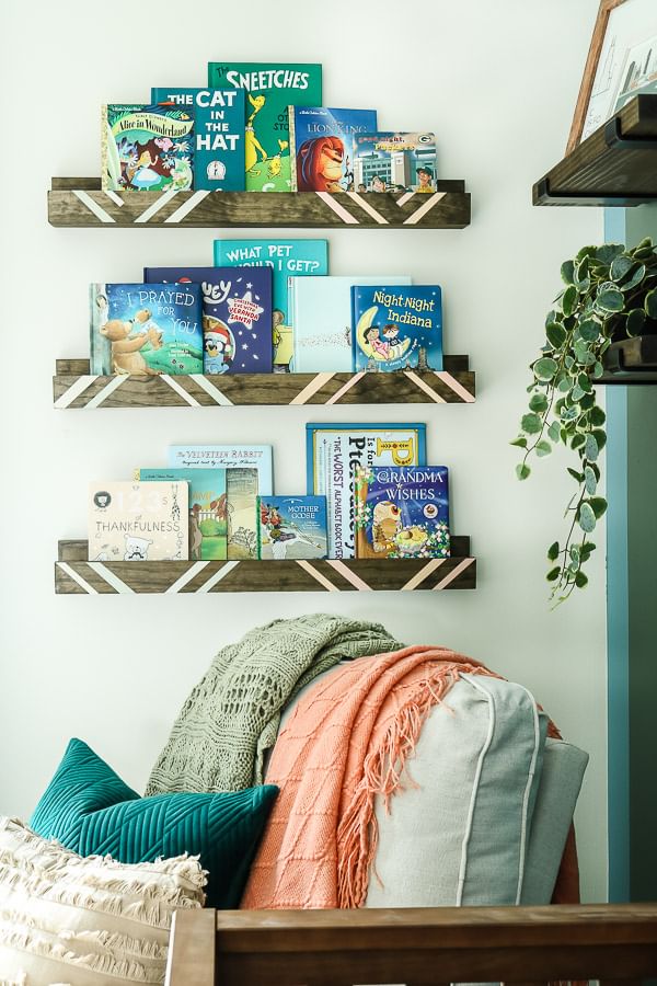 Wooden Book Shelves with crisp white painted lines, hung on a wall. There are children's books on the shelves and there is a cozy chair with blankets on the cushions.