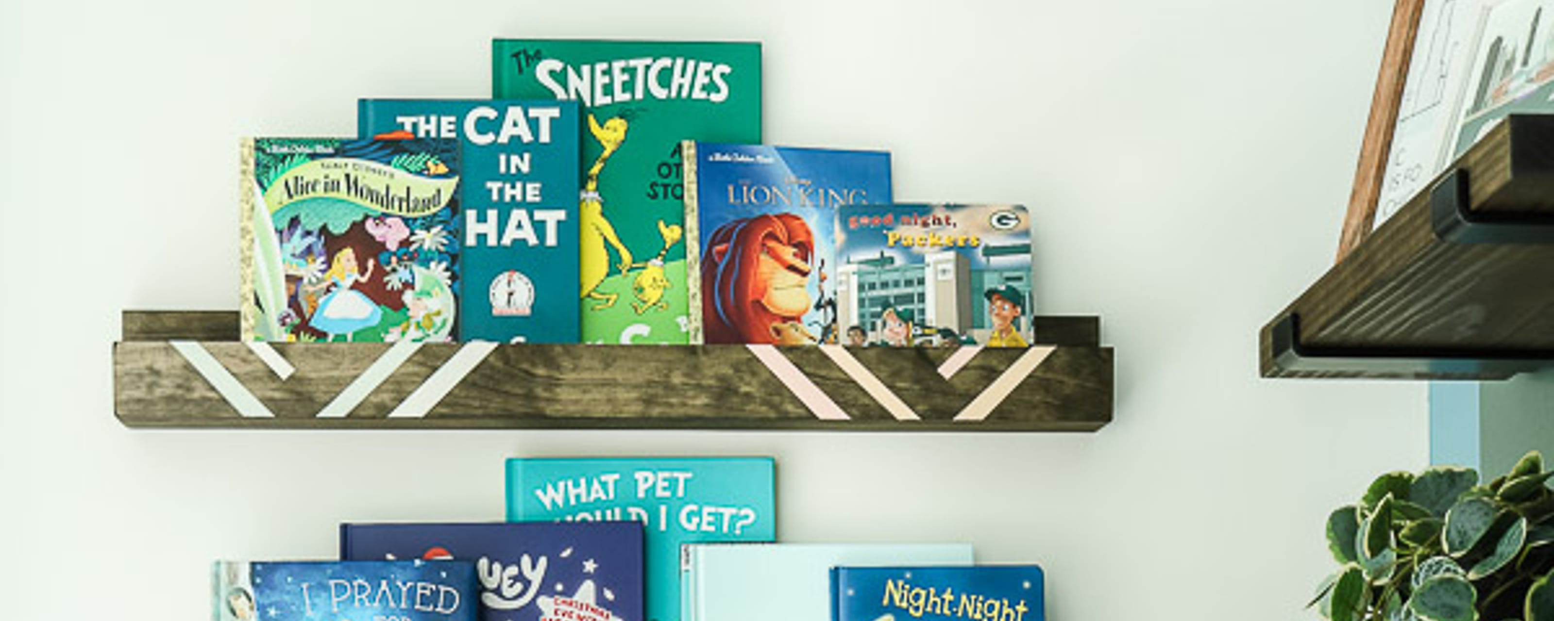 Wooden Book Shelves with crisp white painted lines, hung on a wall. There are children's books on the shelves and there is a cozy chair with blankets on the cushions.