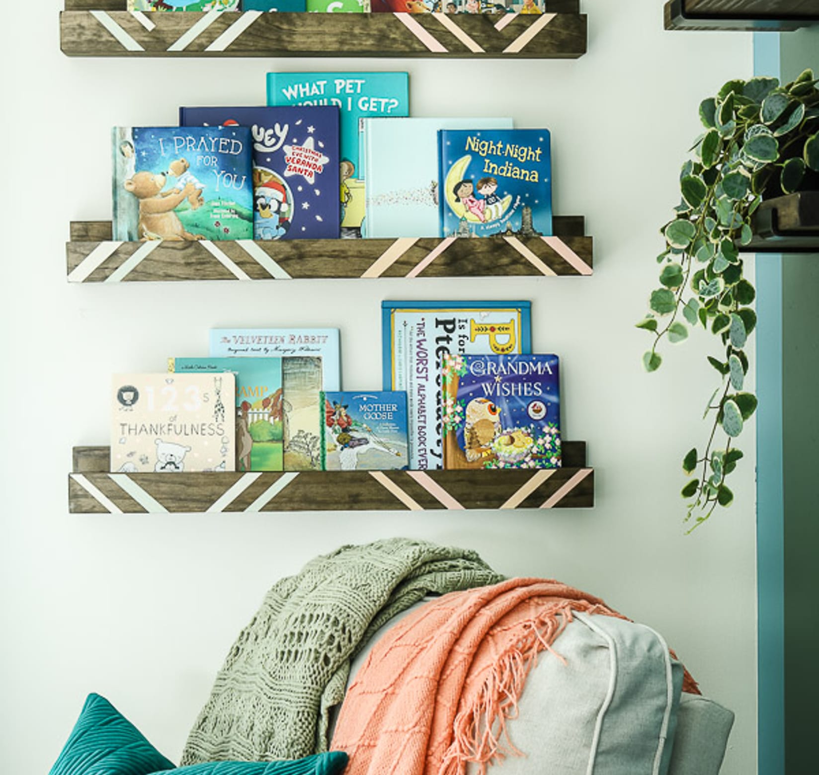 Wooden Book Shelves with crisp white painted lines, hung on a wall. There are children's books on the shelves and there is a cozy chair with blankets on the cushions.