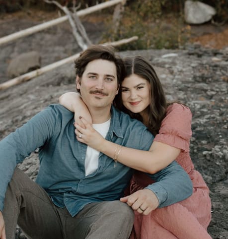 A couple sits on rocky ground, the woman in a pink dress with her arm around the man in a blue shirt. They smile warmly, conveying a sense of joy.