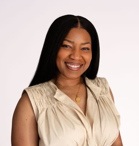 Smiling woman with long dark hair wearing a beige sleeveless blouse and a necklace, set against a plain white background. The tone is warm and friendly.