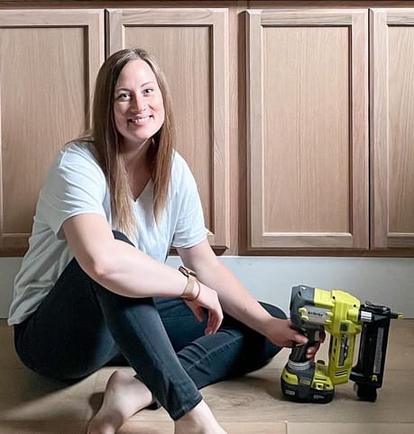 Smiling woman sits barefoot on wooden floor, holding a yellow nail gun. She is in front of wooden kitchen cabinets, creating a relaxed, DIY atmosphere.