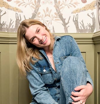A smiling woman in a denim jumpsuit sits against a wall with a nature-themed wallpaper. She holds her knee, exuding a relaxed and cheerful vibe.