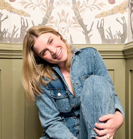 A smiling woman in a denim jumpsuit sits against a wall with a nature-themed wallpaper. She holds her knee, exuding a relaxed and cheerful vibe.