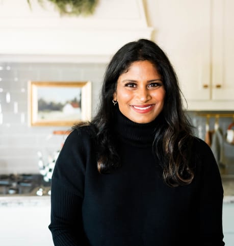 Smiling woman in a black sweater stands in a cozy kitchen, holding a cute animal-shaped mug. A wreath and framed picture adorn the background.