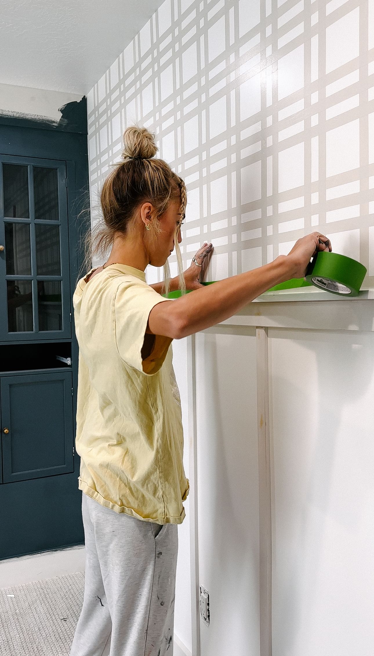 A person wearing a yellow shirt and gray pants tapes a plaid-patterned wall. The room has a calm, DIY atmosphere with a blue cabinet nearby.