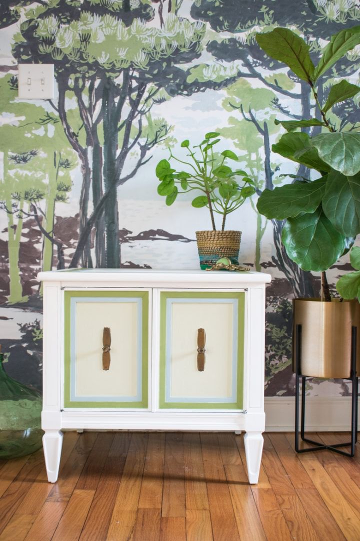 A small white end table with 2 doors lined in green with brown handles. The back wall has a mural with trees and different plants