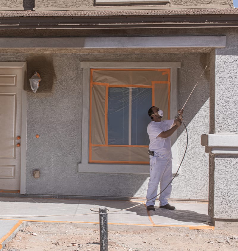 House with a man painting the outside. The windows, cement, and garage door are taped off with FrogTape Pro Grade Orange Painter's Tape