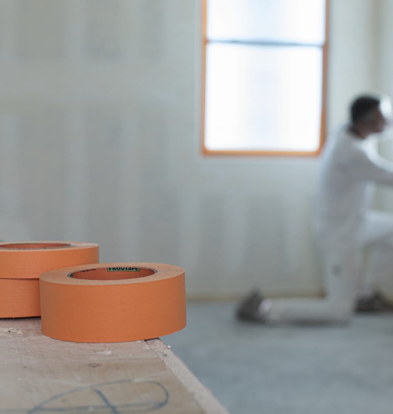 Three rolls of FrogTape Pro Grade Orange Painter's Tape with a man masking a paint site's windows in the background.