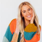Smiling woman with long blonde hair wearing a colorful, chunky knit sweater in orange, teal, and mustard. She rests her head on her hand.