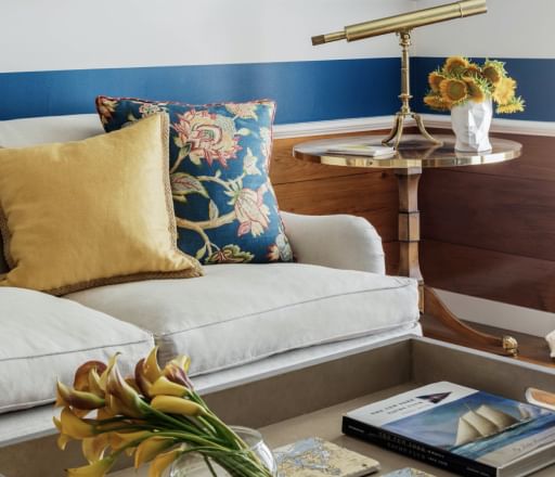 Cream couch with gold throw pillow and flower patterned throw pillow. Coffee table with flowers, coasters, and a book, and end table with flowers and a golden telescope. Back wall is white with thick blue stripes.
