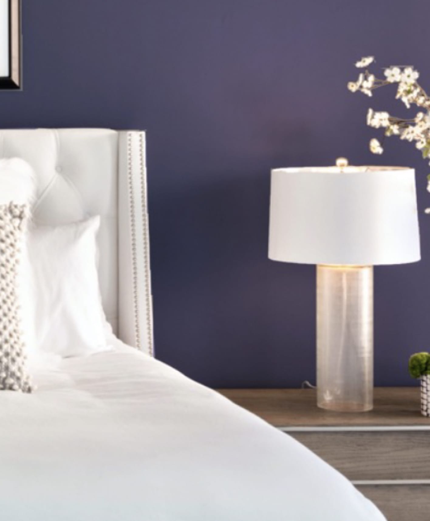 Bedroom with calming purple wall. Bed with White sheets, pillows, and headboard. Nightstand with lamp, succulent, and vase with flowers.