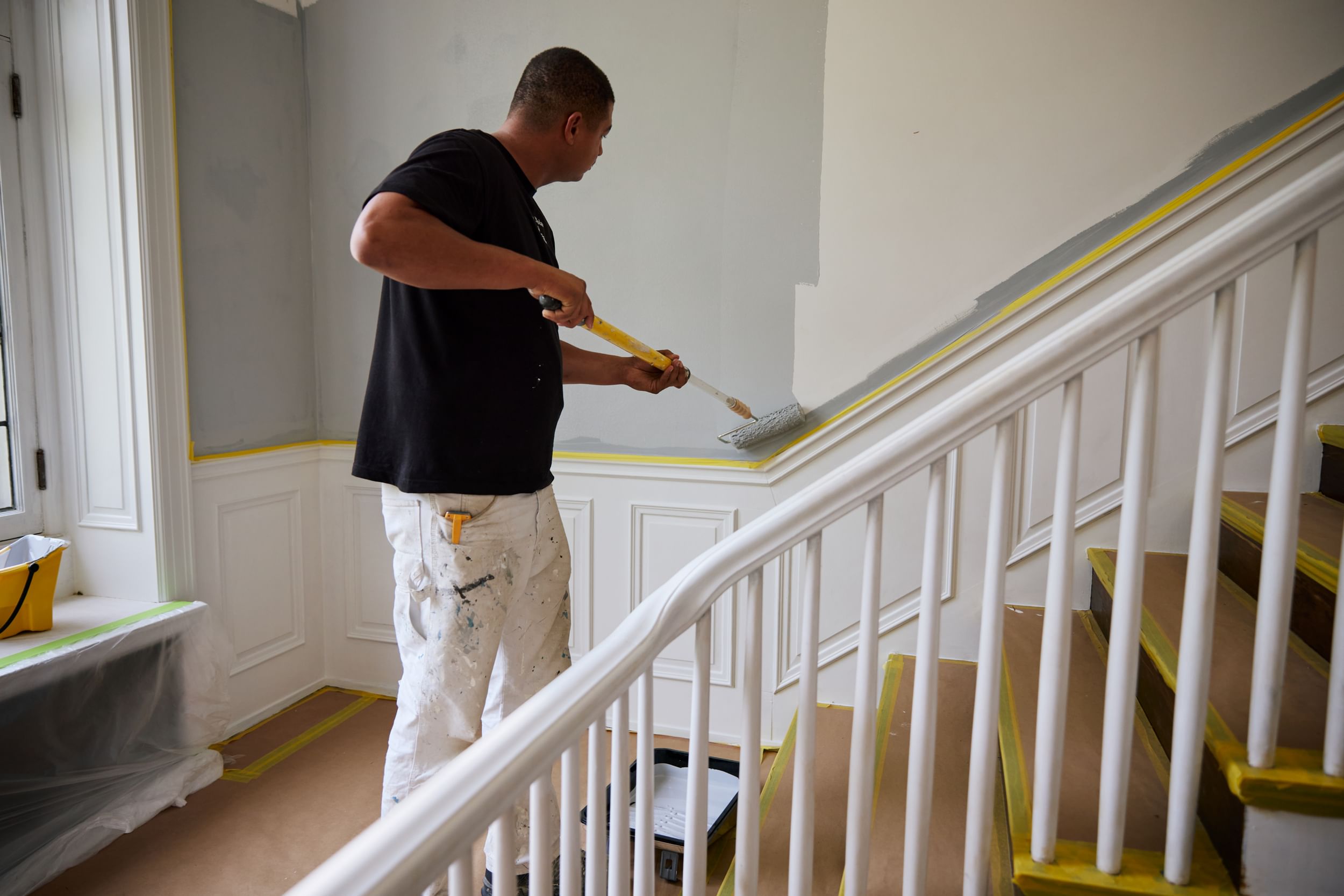 A man using a paint roller to paint the wall going up a staircase gray. Yellow FrogTape Delicate Surface Painter's Tape is used on the trim and steps