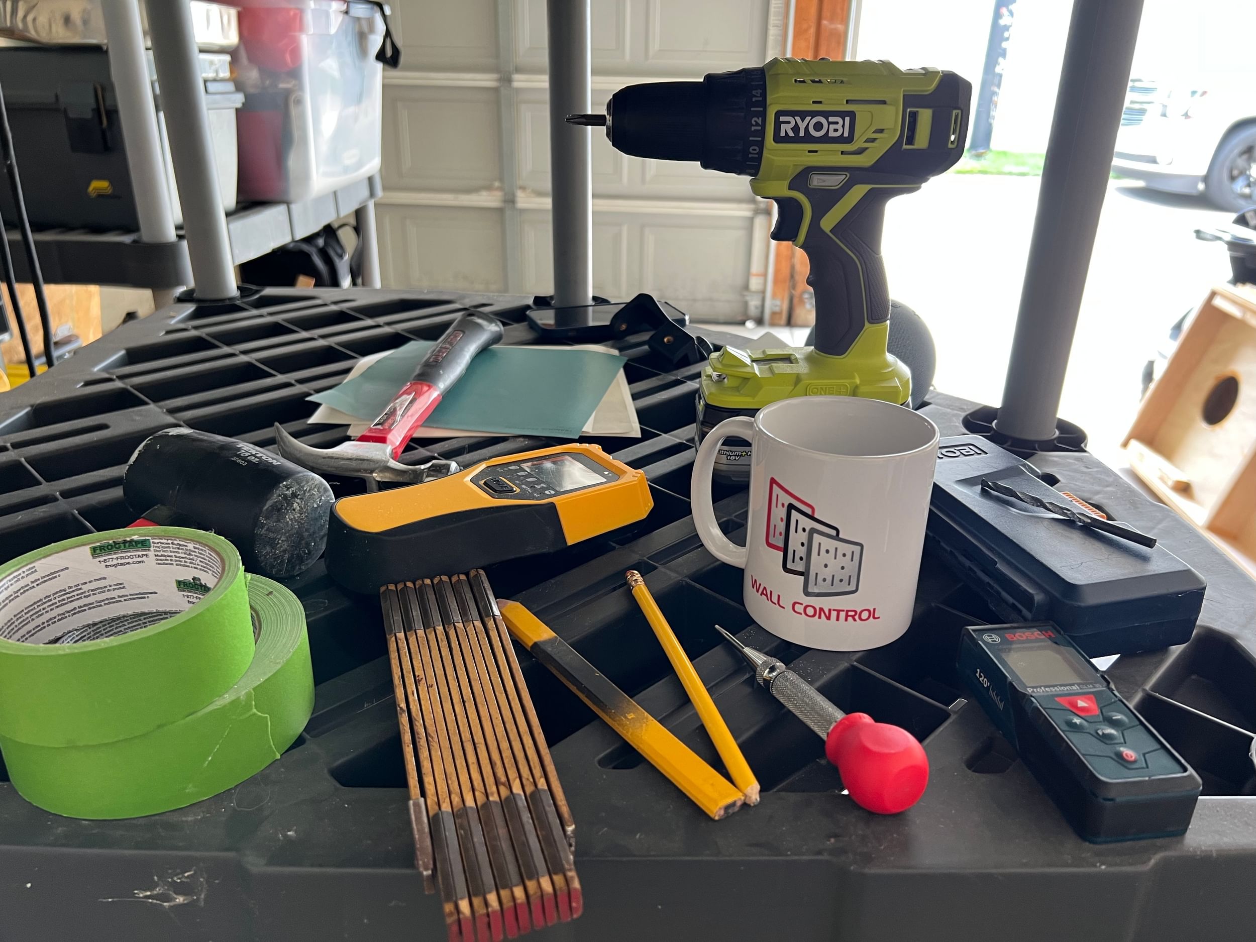 Black Shelf with various different tools. (2 rolls of FrogTape Multi-Surface Painter's tape, pencils, a mallet, remote, coffee mut, drill, hammer, drill bits, and a folder with paper)
