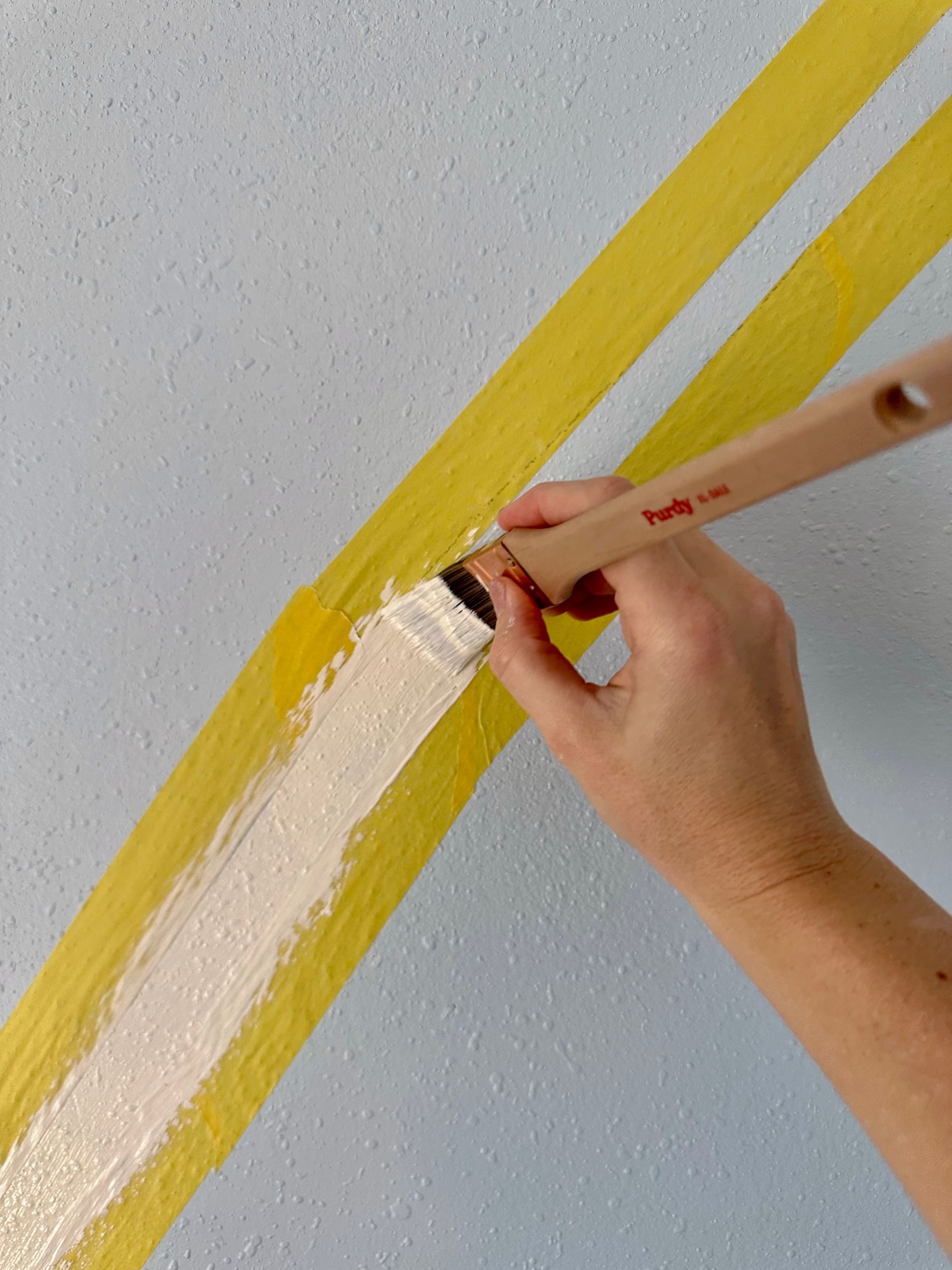 White paint being applied in between two lines of Yellow Delicate Surface Painter's Tape on a blue wall with a paint brush