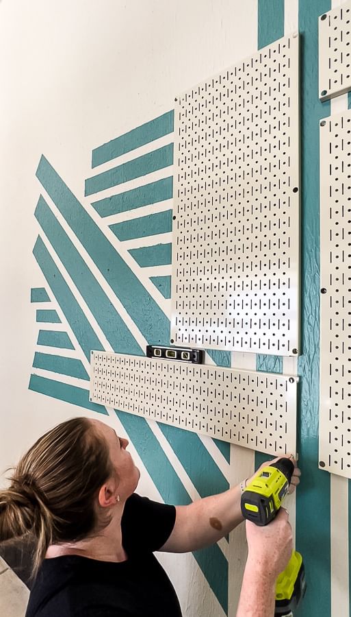 Woman drilling white pegboard sheets onto painted blue garage wall design.