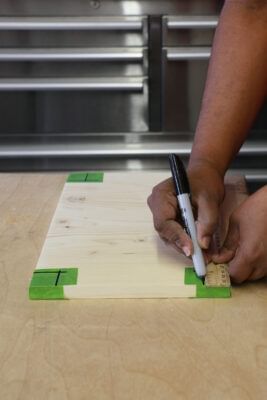Green FrogTape Multi-Surface Painter's Tape on the four corners of a board. Ther is a person marking off notches with a marker.