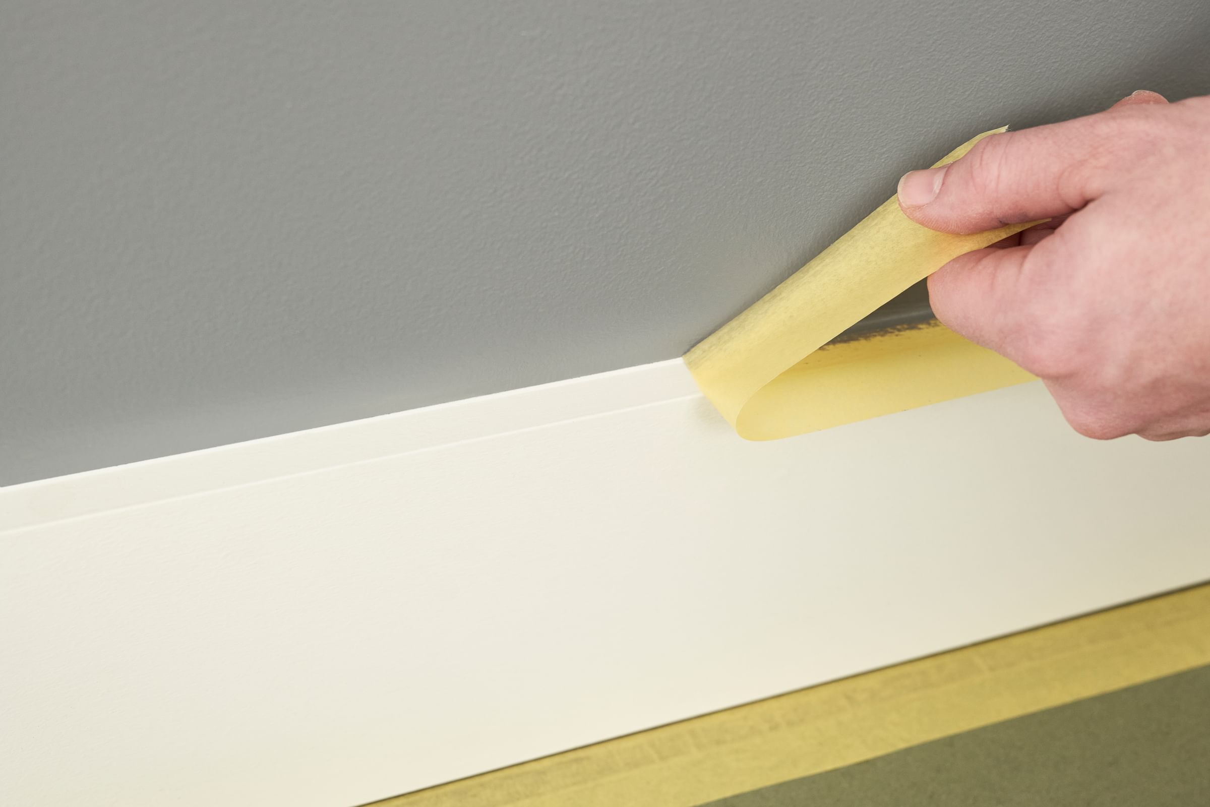 A hand pulling Yellow FrogTape Delicate Surface Painter's Tape off of baseboard trim to reveal a crisp paint line between the white baseboard and gray wall