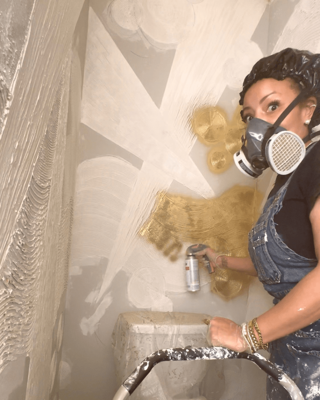Woman in overalls uses gold spray paint and wears a respirator mask in a bathroom. Abstract designs in white and gold cover the walls, conveying creativity.