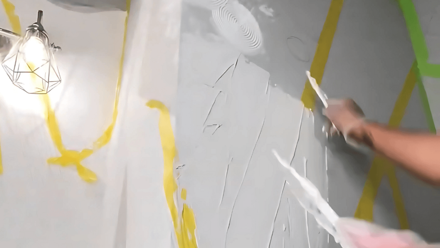 Person applying plaster to a wall with a trowel, featuring a geometric design with yellow tape. A modern, lit lamp hangs on the left.
