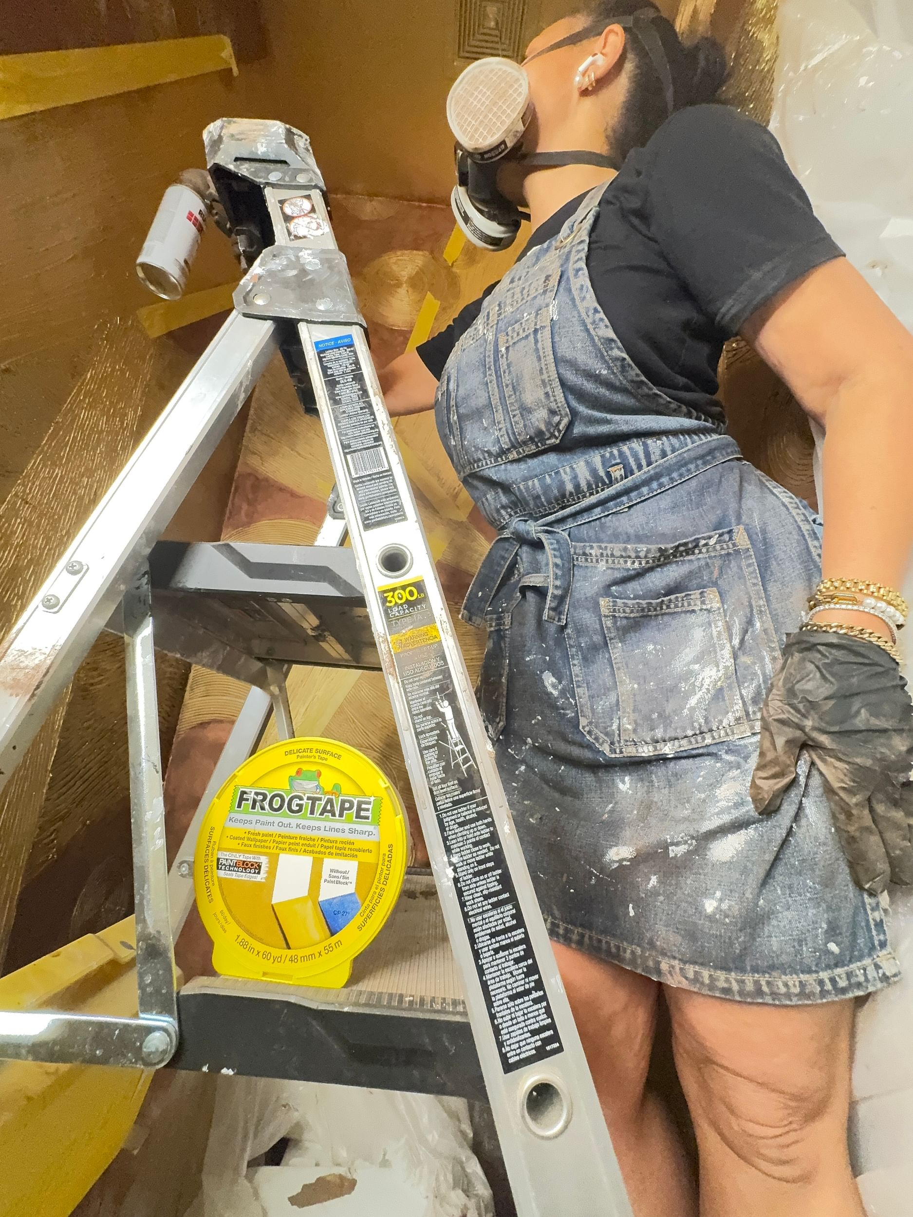 A person in a denim apron stands on a ladder, wearing a respirator mask and gloves, painting. A roll of FrogTape Delicate is visible on the ladder.