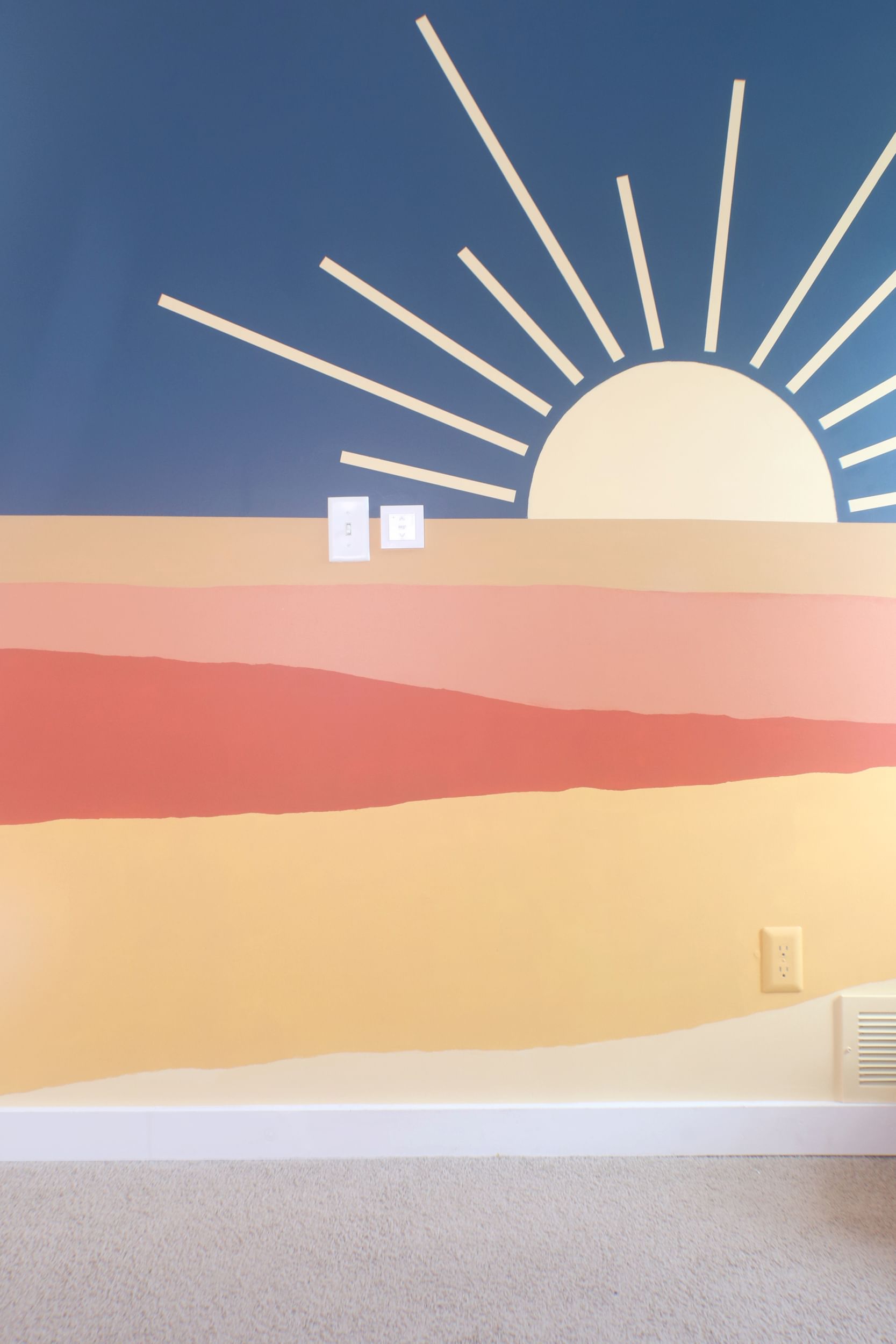 Sunset Wall Mural, (Top to bottom) Blue backdrop with yellow sun with large rays poking up from behind yellow orange layer. Salmon colored layer, organge layer, gold layer, light yellow layer, white baseboard trim, beige carpet.
