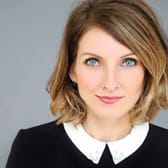 A woman with short, wavy hair wearing a black top with a white collar poses against a gray background, conveying a professional and confident demeanor.