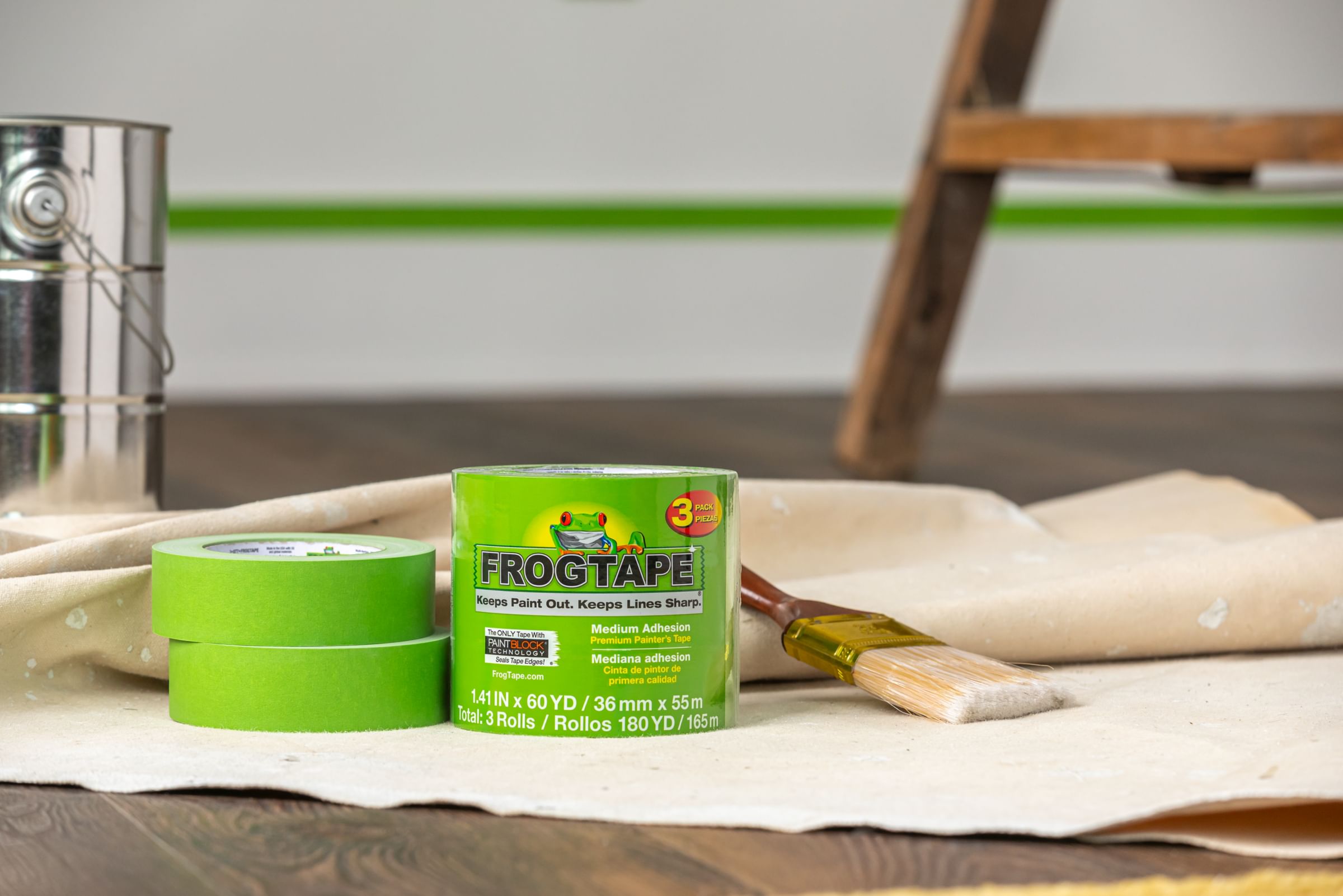 Green FrogTape Multi-Surface Painter's Tape 3 Pack on a paint drop cloth. On the left there are two rolls stacked on top on one another, and on the right there is a paintbrush