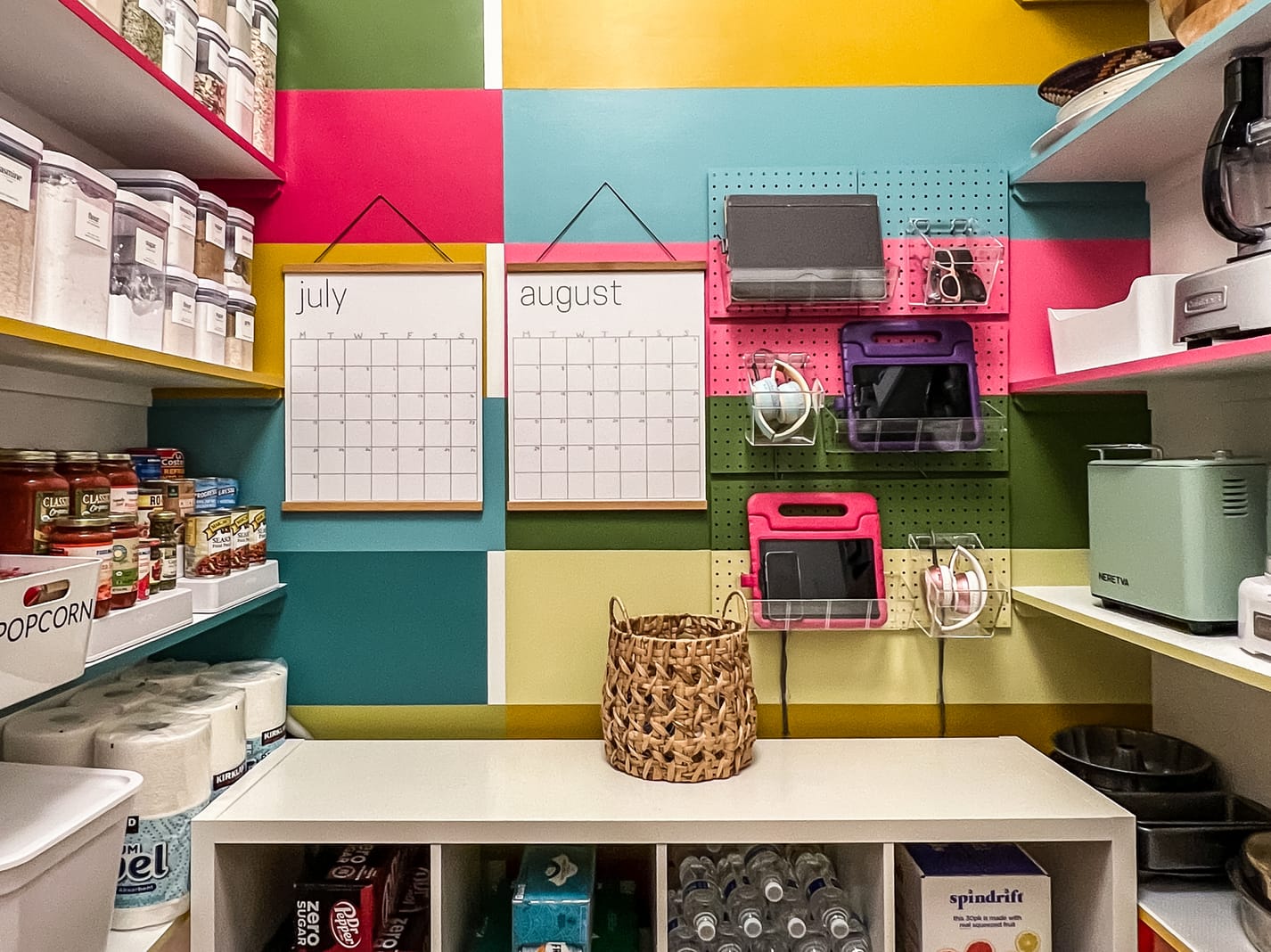 Colorful pantry with organized shelves and storage. Wall calendars for July and August are centered among bright squares. Neat, tidy, and vibrant setting.