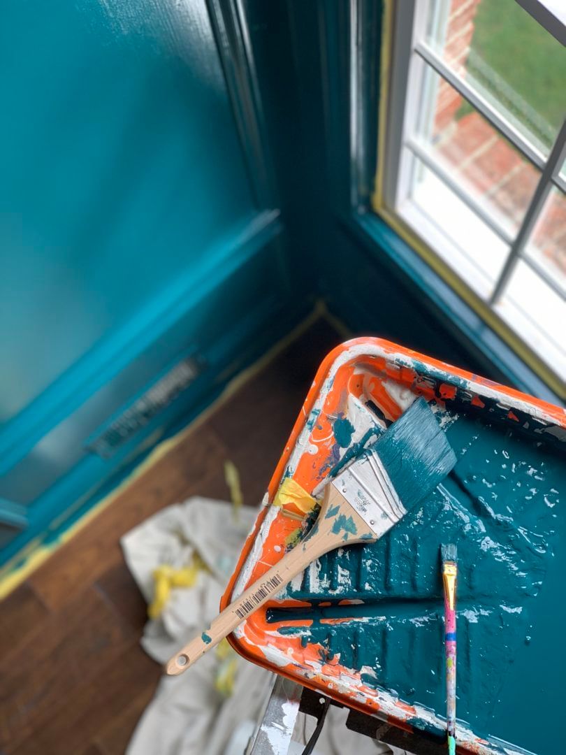 Wall being painted blue. There is a paint tray with a large and small paint brush, both covered in blue paint.