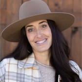 Smiling woman with long dark hair wearing a wide-brimmed hat and plaid jacket. She stands against a wooden backdrop, exuding warmth and friendliness.