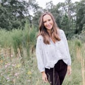 A woman stands in a lush, green field smiling warmly, wearing a white blouse and dark pants. Wildflowers bloom nearby under an overcast sky.