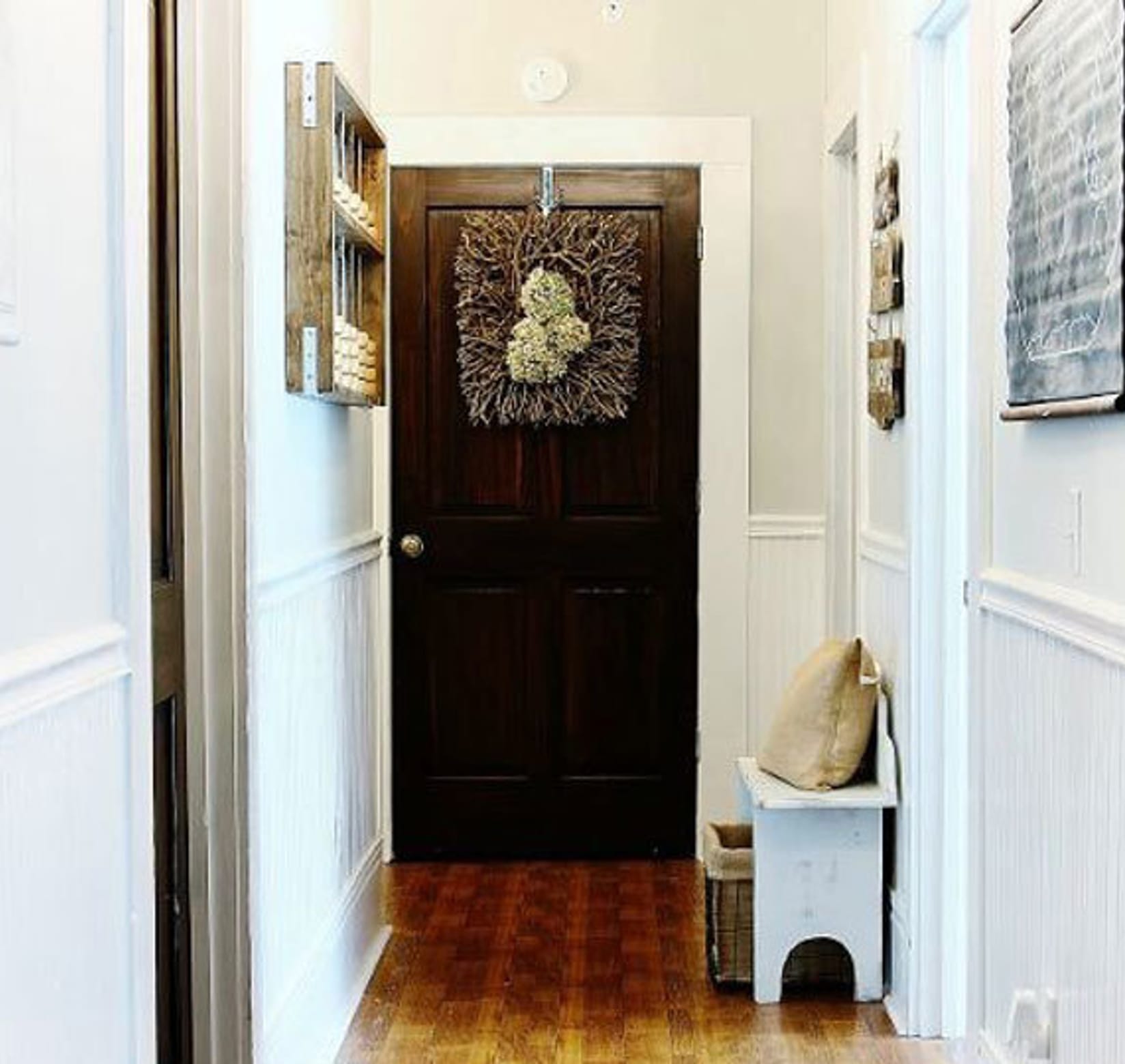 Entryway hallway with a black door at the end. The walls are white with white wainscotting and art on the walls. There is a small bench on the right near the door