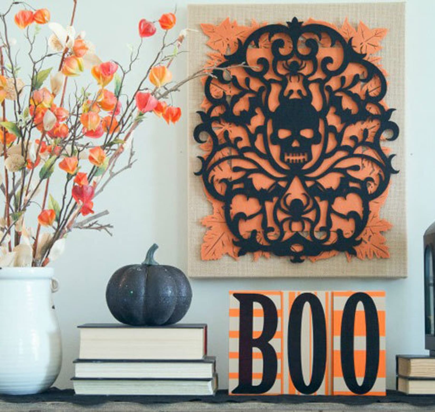 A white vase with flowers, a stack of three books with a black pumpkin on top, BOO Blocks, a stack of two books with a black lantern on top, and on the wall in the center a piece of artwork with a skull in the center