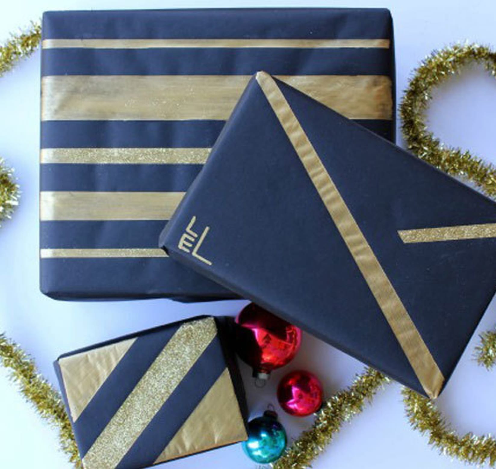 Three black boxes with linear gold designs. There is gold garland on either side of the presents and three bulb ornaments (Two red and one green)