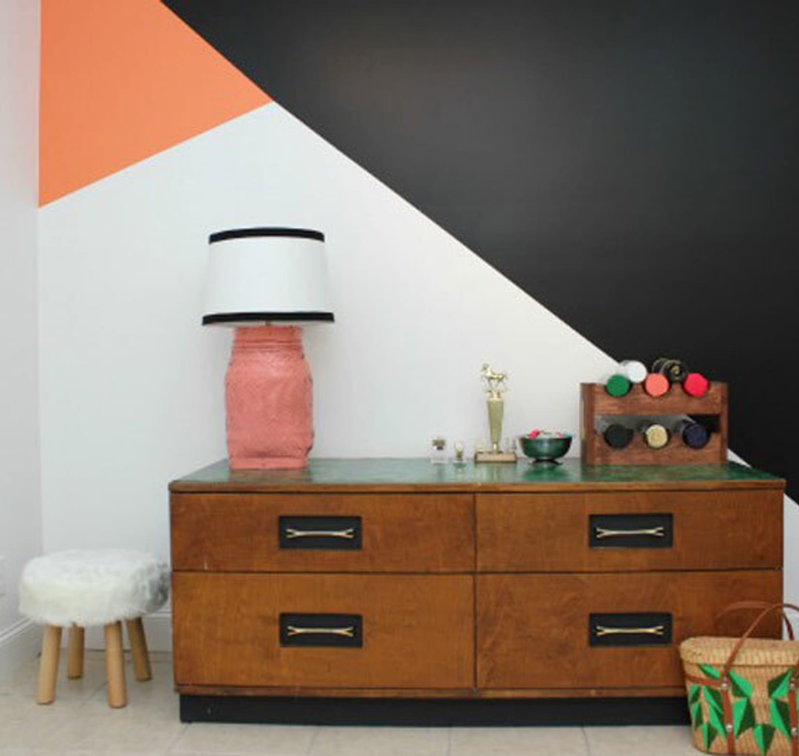 Finished geometric wall mural, orange, white, and black. In front is a small wooden drawers with a pink lamp and other small items atop. There is a small white stool on the left of the drawers and a basket on the right.