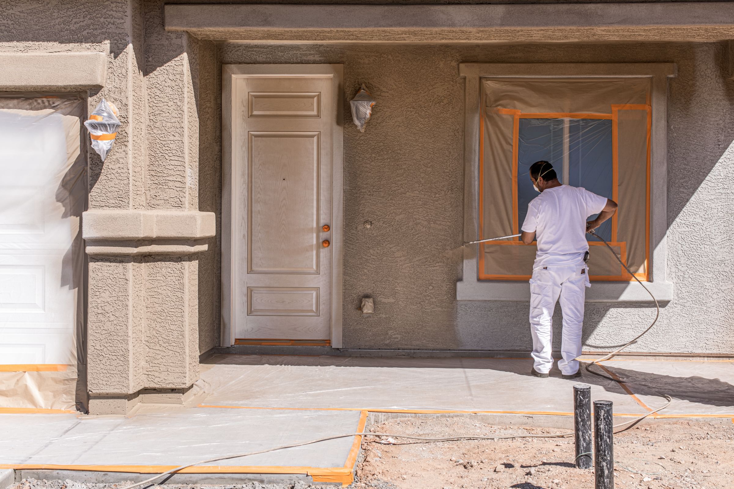 A painter spraying a house with beige paint. FrogTape Pro Grade Orange Painter's Tape is used to help cover the windows and other areas that need to be protected from the paint