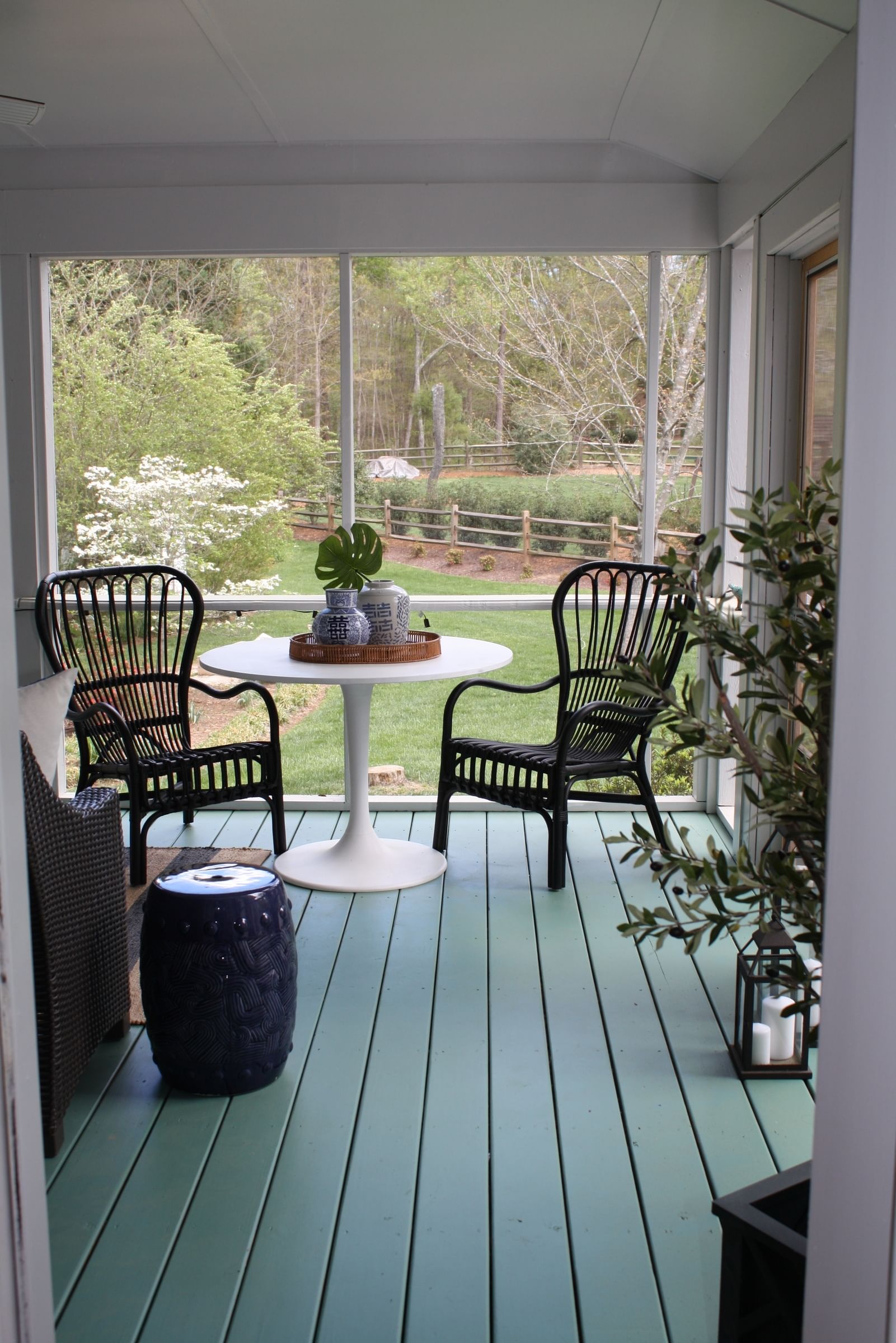 Covered porch with two black chairs and round white table on blue-green flooring. Plants and decorative jars adorn the table, overlooking a lush garden.