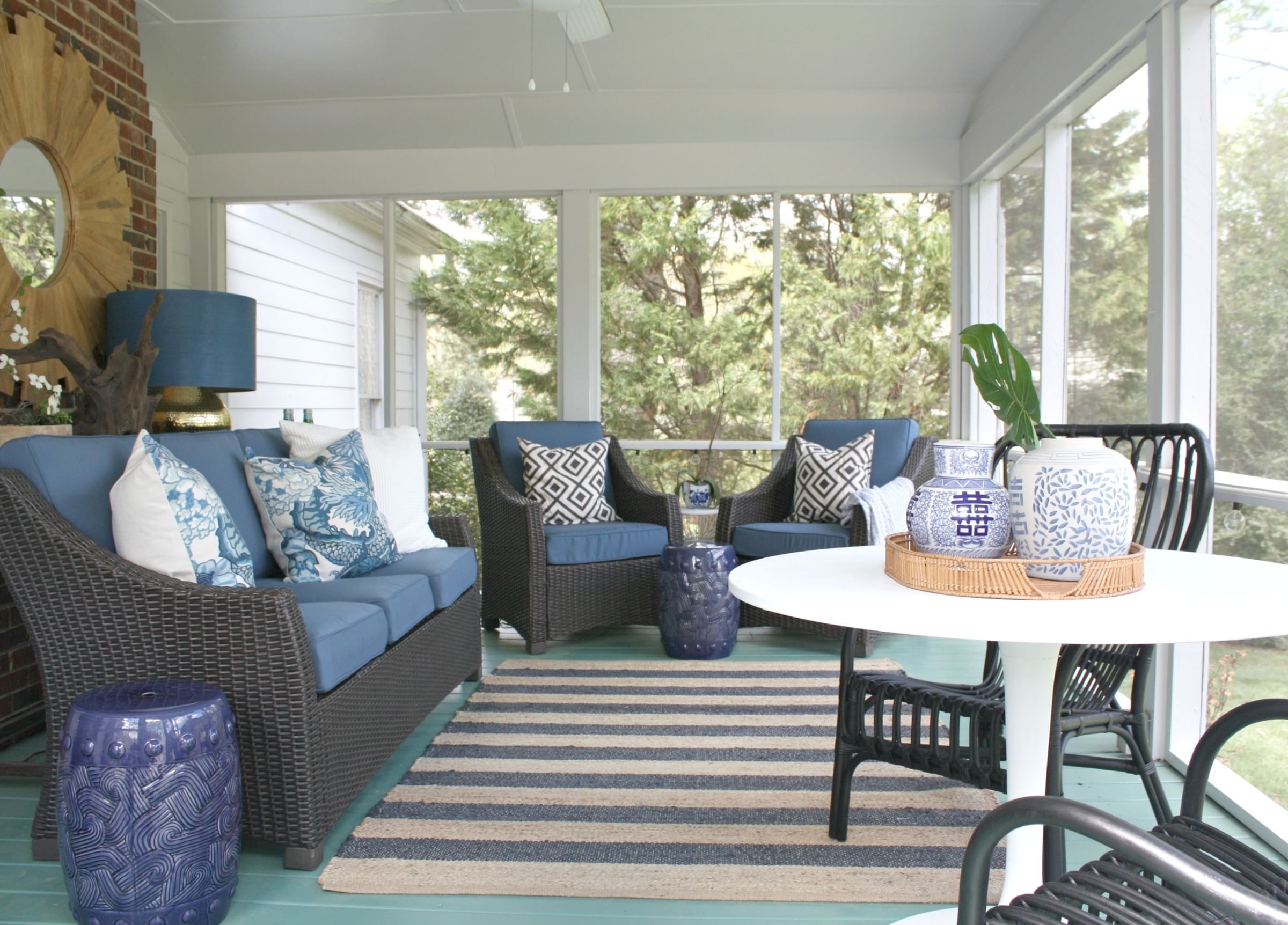 Cozy sunroom with wicker furniture featuring blue cushions and patterned pillows, a striped rug, and decorative vases on a white table. Bright and tranquil.