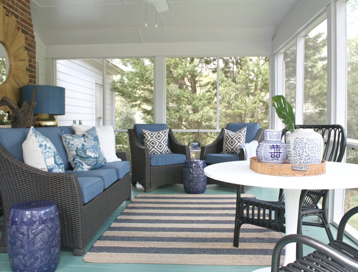 Cozy sunroom with wicker furniture featuring blue cushions and patterned pillows, a striped rug, and decorative vases on a white table. Bright and tranquil.