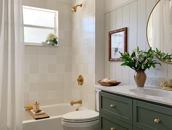Bright bathroom with white tiles and gold fixtures. A round mirror hangs above a green vanity with flowers. Cozy, inviting, and elegant atmosphere.